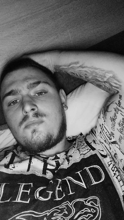 This black and white image displays an adult male positioned indoors in the city of Senec, Slovakia. The subject is lying down, with his head resting on a white pillow. He has short, dark hair, a trimmed beard, and a mustache, with his eyes open and directed towards the viewer. His facial expression is neutral. He is wearing a dark t-shirt with prominent light-colored graphics on the front, partially displaying the word "LEGEND." Visible tattoos include a full sleeve on his right arm and text on his left bicep, partially legible as "authority for me my father!". His right arm is bent with the hand positioned behind his head. The immediate background shows a plain, textured surface behind the pillow, likely a wall or headboard.