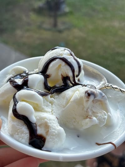 A close-up shot shows a white bowl filled with scoops of vanilla ice cream drizzled with chocolate syrup. A silver spoon is partially submerged in the ice cream, resting on the rim of the bowl. The background is a blurred green and brown landscape, suggesting an outdoor setting. The lighting indicates it is daytime, possibly outdoors due to the natural light and blurred greenery. The focus is on the ice cream, highlighting its creamy texture and the glossy chocolate sauce. The overall impression is one of a refreshing dessert on a pleasant day. There is no visible text in the image.