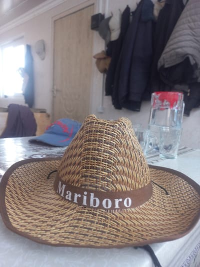 A straw cowboy hat with a brown band that reads "Mariboro" in white letters is the main subject of the image. The hat is placed on a table in the foreground. In the background, there is a blue baseball cap with red lettering, a purple garment draped over a chair, and a stack of clear glasses with a red-rimmed glass on top. Clothes hang on a rack behind the table. The scene appears to be indoors, possibly in a residential setting, with a window and a door visible in the background. The lighting suggests it is daytime. There are no people visible in the image. The location in the city of Şəfalı, Azerbaijan is not discernible from the image itself.