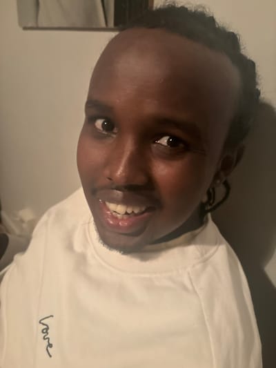 A medium close-up, rotated portrait shows a young person with dark skin and short, braided hair. They are looking directly at the camera with a bright, engaging smile, revealing their teeth. Their eyes are dark and reflective of the light source. The person is wearing a white, long-sleeved crew-neck shirt. On the front of the shirt, near the bottom right, the word "love" is embroidered in black cursive script. The background is a plain, light-colored wall, suggesting an indoor setting. The lighting is soft but bright, highlighting the person's face. The image appears to be taken at an angle, giving the impression of a tilted head or camera. The subject's expression conveys happiness and warmth.