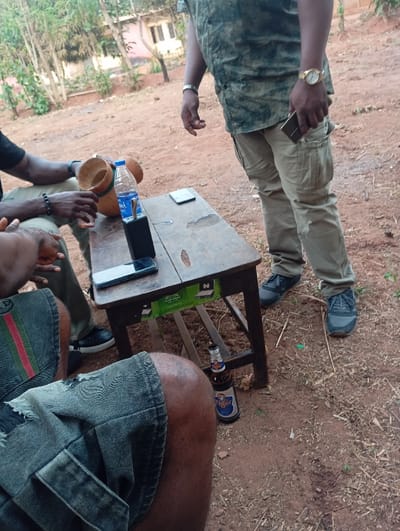 Four individuals are gathered around a small wooden table. On the table are two cell phones, a black power bank, a water bottle, and a calabash. One person is standing, wearing a camouflage shirt, khaki pants, and a gold watch. Under the table, a bottle of Tiger beer sits on the ground. The setting appears to be outdoors, with a building visible in the background. Two men sit at the table, one in gray shorts with a stripe and the other in olive pants.