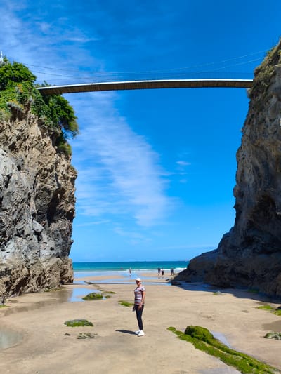 The image depicts a scene at Towan Beach in Newquay, United Kingdom. A suspension bridge spans across a gap between two rocky cliffs, with lush green vegetation growing on the left cliff. The beach is sandy with patches of water and green algae-covered rocks. A woman wearing a striped t-shirt, dark pants, and a cap stands on the sand in the foreground, facing towards the sea. In the background, the blue ocean meets the horizon under a clear blue sky with scattered white clouds. Several people are visible in the distance walking on the beach and in the water. The overall atmosphere is sunny and summery.
