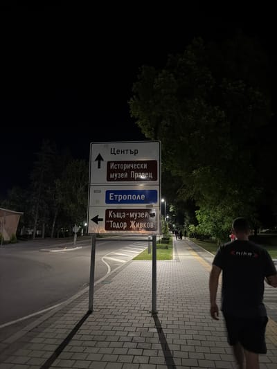 The image depicts a nighttime scene in the city of Pravets, Bulgaria, featuring a street, a sidewalk, trees, and a directional signpost. The signpost displays directions to "Център" (Center), "Исторически музей Правец" (Historical Museum Pravets), "Етрополе" (Etropole), and "Къща-музей 'Тодор Живков'" (House-Museum 'Todor Zhivkov'). A person is walking away from the camera on the sidewalk, wearing a dark t-shirt with a "Nike" logo. Streetlights illuminate the sidewalk and parts of the street. The environment appears to be a public area with pedestrian pathways and road infrastructure.