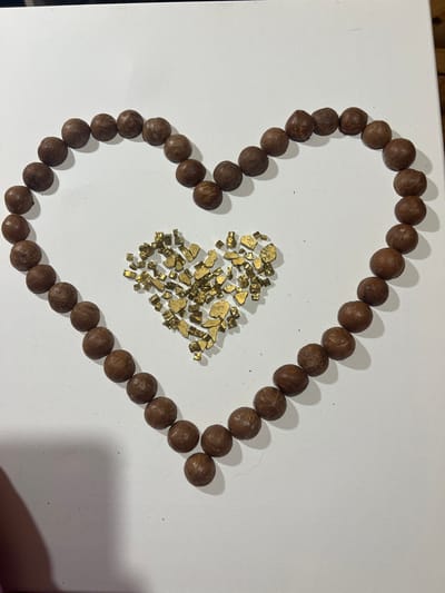 The image shows a heart shape formed by brown, spherical objects resembling nuts arranged on a white surface, likely a canvas or board. Inside the heart shape is a pile of small, irregularly shaped golden objects. The setting appears to be indoors, on a flat surface. The image was taken in Ильинская Поляна Снт, Russia.