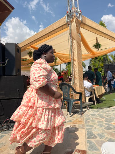 This image captures an outdoor event in Kumasi, Ghana, under clear blue skies with scattered white clouds. The primary subject is a dark-skinned woman in the foreground, positioned in profile facing right. She wears a cream-colored dress with a red floral pattern, featuring puff sleeves and multiple ruffled tiers. Her dark hair is styled in braids pulled back, and she has a light-colored earring on her visible ear. She is wearing light-colored open-toed shoes, and her body is partially shadowed.
To the left of the woman, two large black speakers are stacked. In the midground, a large event canopy structure with gold/tan fabric drapes and a metal truss system is prominent. Hanging decorative greenery is visible beneath the canopy. Chains descend from a vertical support beam of the canopy.
Under the canopy and extending into the background, multiple individuals are present. Several are seated at tables covered with light-colored cloths; one man in a white shirt is partially visible sitting at a table on the right. Other individuals are standing, including men in dark green attire and a man in a light blue shirt. A dark gray plastic chair is positioned near a canopy support. The ground in the foreground consists of flagstone paving, transitioning to artificial green turf or grass further back. Green foliage and trees are visible in the background, along with a dark metal gate. The scene is brightly lit by sunlight, casting shadows.