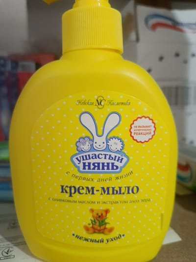 This media file features a bright yellow plastic bottle of "Ushastiy Nyan" (Ушастый Нянь) cream soap, designed for babies. The bottle has a pump dispenser at the top. The label is yellow with white polka dots and features illustrations of a cartoon bunny and a teddy bear.
Prominently displayed on the label are several lines of text in Russian. At the top, it reads "Невская NC Косметика" (Nevskaya NC Cosmetics). Below that, in a blue banner, is the brand name "Ушастый Нянь" (Ushastiy Nyan). Underneath, it states "с первых дней жизни" (from the first days of life). The main product name, "крем-мыло" (cream soap), is written in large blue letters. Further text below mentions "с оливковым маслом и экстрактом алоэ вера" (with olive oil and aloe vera extract). At the bottom, the phrase "нежный уход" (gentle care) is visible.
To the right of the bunny illustration, a red, scalloped circular badge contains text in Russian that translates to "DOES NOT CAUSE ALLERGIC REACTIONS". The overall impression is of a gentle, child-friendly product. The background is blurred but appears to be a retail setting with shelves and other products, suggesting the item is for sale. There are no discernible cues for time of day or weather. The location context indicates Berdiansk, Ukraine.