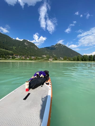 This media file depicts a serene scene on a lake in Lärchenwiese, Austria. The perspective is from a stand-up paddleboard, showing the front of the board and the surrounding environment. The water is a striking turquoise-green, with gentle ripples indicating calm conditions.

Dominating the background are lush green mountains covered in dense forest, with patches of snow visible on the higher peaks. A small village or cluster of houses can be seen nestled along the shoreline, surrounded by reeds and trees. The sky is a vibrant blue, punctuated by fluffy white clouds, suggesting a clear and pleasant day, likely late morning or early afternoon given the bright sunlight casting shadows.

On the paddleboard, a black bag with purple accents is secured, along with a can of Budweiser. The paddleboard itself is white with orange detailing and a textured grey surface. The overall impression is one of tranquility and outdoor recreation. The scene evokes a sense of peace and enjoyment, likely during a summer day's paddle.