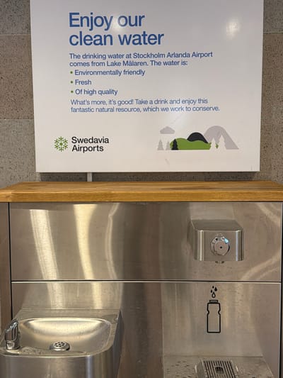 The image shows a public water fountain at Stockholm-Arlanda Airport, Sweden. A sign above the fountain states, "Enjoy our clean water." It further explains that the drinking water comes from Lake Mälaren and is environmentally friendly, fresh, and of high quality. The sign also features the Swedavia Airports logo and a graphic of mountains and trees. The water fountain itself is made of stainless steel and has a visible water spout on the left and a bottle filling station with a graphic of a bottle and dripping water on the right. The surrounding environment appears to be an indoor public space, likely an airport terminal, with textured walls.