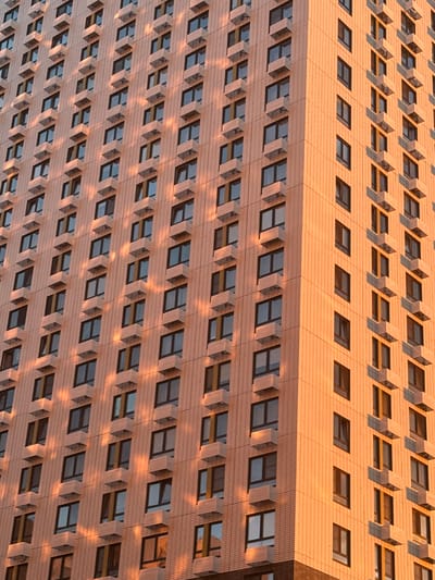 This is a close-up, high-angle, eye-level shot of a modern building facade, likely an office building or apartment complex, illuminated by the warm, golden light of either sunrise or sunset. The building is constructed with light-colored, textured panels, possibly brick or concrete, arranged in a grid pattern. Rows of rectangular windows are recessed and feature protruding, angled shades that cast strong, geometric shadows across the facade. The windows themselves appear dark, reflecting the sky or interior lighting, with some hints of blue or orange light reflecting from the sky. The repeating pattern of windows and shades creates a strong sense of rhythm and order. The sun's low angle casts long, dramatic shadows, highlighting the architectural details and adding depth to the image. The overall impression is one of architectural uniformity and the play of light and shadow on a large, man-made structure. There are no visible people, text, or specific identifiable landmarks that would pinpoint an exact location beyond the general context of a modern urban or suburban environment.