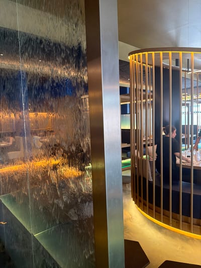 The image shows an interior scene with a layered perspective. In the foreground, a reflective surface with the distorted reflection of an overhead structure. This structure appears to be a partially enclosed dining area with horizontal wooden slats. People are seated within this area.

The environment appears modern, with sleek lines and muted tones. The image was taken in Torreforta, Spain.
