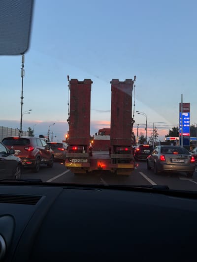 In Troitsk, Russia, at dusk, a line of vehicles is stuck in traffic. The perspective is from inside a car, looking forward through the windshield. Ahead, a large, red, two-part trailer rig dominates the view. Its rear lights are illuminated. To the right of the trailer, a fuel station sign displays prices: 63.65, 72.89, and 98.38. Several cars are visible in the traffic jam, including a red SUV and a silver sedan. The sky is a pale blue with hints of pink and orange from the setting sun. Streetlights and power poles are visible along the roadside, along with a dense line of trees in the background. A partial view of the car's dashboard is in the foreground, along with a portion of the windshield wiper.