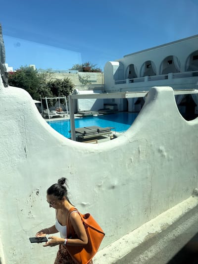 In the city of Thira Municipal Unit, Greece, a woman walks in the foreground holding a smartphone, looking down at it. She has her hair in a bun, wears sunglasses, a white tank top, and a patterned skirt. She carries an orange tote bag over her right shoulder. Behind her, a swimming pool with lounge chairs and umbrellas is visible, with a resort-style building in the background. The scene is outdoors on a sunny day, reflected in a window.
