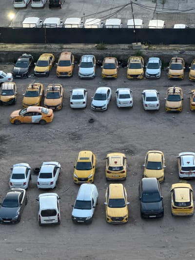 The image is an aerial view of a parking lot filled with numerous yellow and white cars, many of which appear to be taxis. The cars are parked in neat rows on a gravel surface. In the background, there are several white modular structures lined up against a dark fence. A few trees and bushes are visible near the fence. The overall impression is of an organized fleet of vehicles, possibly awaiting dispatch or maintenance. The scene suggests a functional, utilitarian space rather than a residential or commercial area.