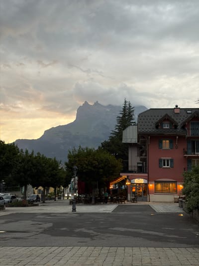 The image depicts a street scene in Saint-Gervais-les-Bains, France, with a prominent mountain peak dominating the background. The sky is overcast with dramatic clouds, tinged with the warm hues of a setting sun on the left.
In the foreground, a wet asphalt road surface is visible, intersected by a pedestrian crossing. The road is bordered by a cobblestone sidewalk.
On the right side of the street, a salmon-pink building with a dark, multi-pitched roof stands out. It has multiple windows, some with dark shutters, and balconies. The ground floor features a cafe with illuminated windows and a lit sign that reads "Café". Warm light spills from the cafe windows and exterior lighting.
Across the street, to the left, a row of trees with dark green foliage lines the sidewalk. Beyond the trees, a line of buildings with yellow facades can be seen, suggesting a typical European townscape. Several cars are parked along the street.
Further down the street, tables and chairs are set up outside another establishment, likely a restaurant or cafe, with red umbrellas providing shade. A few individuals are visible, appearing to be patrons or staff. Vehicles are also present on the road, with headlights illuminated, indicating it is either dusk or a dimly lit area.
The overall atmosphere is serene and slightly moody, with the imposing mountain range serving as a constant backdrop to the urban environment. The image captures a moment of transition, possibly late afternoon or early evening, as the day fades and the town's lights begin to glow.