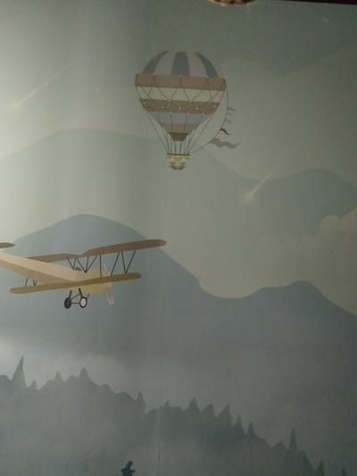 This media file depicts a mural or artwork featuring an airplane and a hot air balloon in a mountainous landscape. The primary subjects are the biplane in the foreground and the hot air balloon in the upper portion of the frame. The background consists of stylized, layered mountains, fading into a soft, blueish-gray sky. Light streaks, possibly representing shooting stars or atmospheric effects, are visible in the sky. The biplane is a light tan color with brown wings and black struts. It has two wheels and a propeller. The hot air balloon has a striped design, with segments of tan and light blue. It is suspended by ropes from a basket. The scene evokes a sense of adventure and flight, with an implied time of day that could be dawn or dusk due to the soft lighting and muted colors. The location cues suggest a fantastical or artistic representation rather than a realistic depiction of a specific place in Kalachinsk, Russia. No text is visible in the media file.