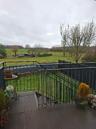 The image displays an elevated view from a residential deck, overlooking a rural landscape in Vallée de l'Ernz, Luxembourg, under an overcast sky.

In the immediate foreground, a dark brown composite deck with parallel grooved planks extends across the bottom of the frame. A black metal railing with vertical balusters outlines the deck and descends via a set of stairs on the right side. To the left, a circular wrought iron table with three legs supports an empty terracotta pot. Below the table, a white spherical garden ornament rests near a small potted green plant, adjacent to a taller, slender potted evergreen. To the right, a large dark grey pot holds a compact, amber-colored topiary-style plant and a smaller green plant with moss. Two light-colored garden gnome-like figures are positioned next to this pot.

Beyond the deck railing, a well-maintained green lawn is visible, enclosed by a dark fence. Further into the midground, a narrow paved road or path runs horizontally, with a light-colored vehicle, possibly a pickup truck or SUV, parked beside a small wooden shed. Numerous bare deciduous trees and some evergreen bushes are scattered across the verdant fields. The background consists of rolling green hills, densely forested at the horizon. The sky is uniformly grey and overcast, with a distinct bright, circular light reflection or anomaly visible near the upper center-left. No people are present in the scene.