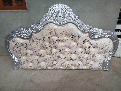 The image displays an ornate, silver-framed headboard with tufted upholstery, positioned upright against a plain wall.
The headboard is the central and dominant object. It features an elaborately carved silver-colored frame that outlines its shape. The top portion of the frame rises into a decorative crest with scrolled and floral-inspired motifs, including a central circular element. The frame then curves downwards along the sides, forming graceful S-curves and further scrolled patterns before straightening out towards the bottom. The silver finish appears slightly metallic and reflective.
The interior panel of the headboard is upholstered in a light cream or off-white fabric. This fabric is deeply tufted with buttons, creating a series of symmetrical indentations across the surface, giving it a plush and luxurious appearance. Overlaid on the light-colored fabric is a distinct, irregular pattern of gold or metallic bronze flecks and splatters, which resembles a distressed or abstract gilded effect, adding texture and visual interest.
The headboard rests on a light gray, possibly concrete, floor or ground surface. The background behind the headboard consists of a plain, light gray wall, which appears to be concrete or stucco. In the upper left corner of the original image, a small section of a dark brown, rectangular object, possibly a wooden window frame or a shelf, is visible against the wall. To the right side of the headboard, in the background, there are some indistinct white objects, possibly boxes or furniture components. One of the cropped images reveals a partial white label or text on one of these background objects, showing the letters "mi" or "im".
No activity is taking place in the image; the headboard appears to be stationary, possibly awaiting installation, storage, or display. The lighting suggests it is daytime, casting soft shadows and highlighting the textures of the headboard. The overall aesthetic of the headboard is opulent and classic, likely intended for a formal bedroom setting.