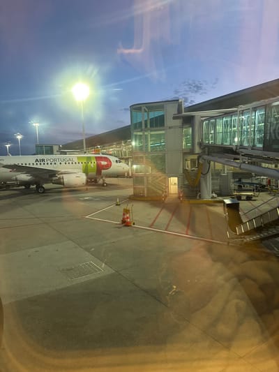 The image presents an airport scene at dusk or night, viewed through a window, possibly from a terminal building or another aircraft. A white Air Portugal aircraft is parked at a gate, connected to a modern glass-fronted terminal building by a jet bridge. The aircraft's fuselage clearly displays "AIR PORTUGAL" in black lettering, and the tail and forward section feature the distinctive green and red 'TAP' logo.

Bright artificial floodlights illuminate the tarmac and the parked plane, casting strong glows, especially from a prominent light pole on the left. The sky is a deep twilight blue with scattered clouds. On the tarmac, white and red painted lines mark pathways and safety zones. Several red and white safety cones are visible near the aircraft's nose, along with a metallic drainage grate. A yellow sign near the jet bridge connection reads "S32," likely indicating a gate or stand number. The window through which the photo is taken shows some reflections and distortions, particularly warm, yellowish light on the right side. The scene depicts typical ground operations at an airport, likely in Moreira, Portugal.