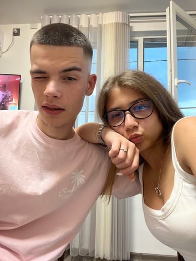 The image depicts two young individuals, a male and a female, posing in a close-up shot within an indoor domestic setting in Karavelovo, Bulgaria.
The male, positioned on the left side of the frame, has short, dark brown hair styled in a buzz cut with faded sides. He possesses fair skin with visible freckles across his nose and cheeks. His left eye is closed in a wink, while his right eye is open, directed towards the viewer. His mouth is slightly agape, revealing the tip of his tongue resting on his upper lip, contributing to a playful expression. He is wearing a light pink crew-neck t-shirt featuring a white graphic on the lower right front, consisting of a palm tree silhouette and cursive text, which appears to read "Santa Mo..." or similar. Faint stubble or peach fuzz is visible along his chin and jawline.
The female, situated on the right, has medium-length, wavy, light brown hair. She is wearing black-rimmed eyeglasses, the lenses of which display a blue reflection, possibly from an anti-glare coating. She has a pouting expression, forming a "kiss face" with her lips, and her eyes are looking directly forward. She wears a white tank top. Her left arm is draped over the male's right shoulder, and her left hand rests against her own cheek or chin. Her fingernails are painted a pale color. Accessories include a silver-toned multi-strand bracelet on her left wrist, a silver-toned ring with a heart-shaped stone on her left ring finger, and a silver-toned necklace with a heart-shaped or intertwined pendant visible at her décolletage.
In the background on the far left, a wall-mounted flat-screen television is visible, displaying an image of a person, likely male, in a white shirt, possibly playing a musical instrument. Above and to the left of the television, a white electrical outlet with a white cable is mounted on the wall. The background on the right features a large window with white frames, partially open inwards on its right side. Light-colored, pleated curtains, possibly sheer or semi-sheer, are drawn across a portion of the window. A clear blue sky is visible outside the window, and a white handle is affixed to the open window section. The overall scene is well-lit, suggesting natural light from the window.