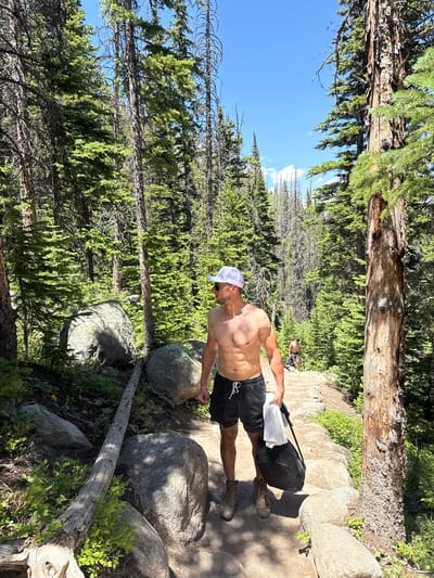 A shirtless man wearing a white baseball cap and black shorts walks up a stone staircase on a hiking trail. He is holding a white t-shirt and a black backpack. The trail is surrounded by a dense forest of pine trees, with large boulders on the side of the path. The sky is clear and blue. The location is likely within Rocky Mountain National Park near Allenspark, Colorado, United States, given the characteristic terrain and vegetation. Another person can be seen walking further up the trail behind the man.