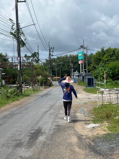 A person wearing a blue hooded jacket, black pants with white stripes, and white boots walks away from the camera on a paved road in Pa Khlok, Thailand. The individual is also wearing a wide-brimmed hat. The road is lined with vegetation and utility poles carrying numerous wires. In the background, there is a blue sign with a white "Lotus" logo and text, and behind that, a building and more trees under a cloudy sky.