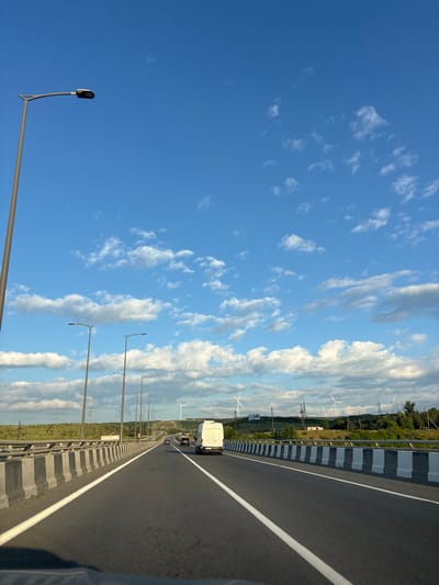 The image depicts a highway scene in the vicinity of Tedotsminda, Georgia, under a bright blue sky dotted with white clouds. A multi-lane asphalt road stretches into the distance, bordered by a concrete barrier with alternating gray and white markings. Several streetlights are visible along the roadside. In the mid-ground, a white recreational vehicle is driving on the highway, followed by other vehicles. The landscape beyond the highway features rolling green hills, some with trees and power lines. In the far distance, several wind turbines are visible, indicating a windy or energy-generating area. The overall scene suggests a sunny day with moderate traffic on a highway passing through a scenic, rural landscape.