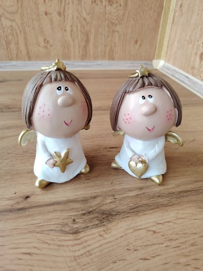 Two small ceramic figurines of angels are positioned side-by-side on a wooden surface. The angels are identical in design, with rounded heads, brown hair, large eyes, pink cheeks with freckles, and small smiles. They are both wearing white robes with gold accents on their wings and feet. The angel on the left is holding a gold star in its hands, while the angel on the right is holding a gold heart. The background consists of a wall with a wood-grain pattern. The lighting suggests an indoor environment. The location is not specified in the image.