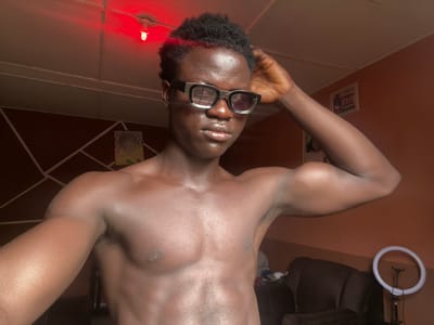 A bare-chested, dark-skinned person with short, curly black hair and wearing black-rimmed sunglasses is lying upside down. The person's arms are raised. A ring light is visible in the upper left corner of the image. A red light is in the lower right corner. The location is indoors. Paintings and a wall decoration are visible in the background. The city and country of Mosafejaw, Nigeria, cannot be definitively determined from the image.