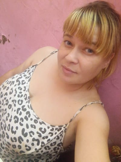 A woman with blonde hair and blue eyes, wearing a white and black leopard print camisole, poses for a selfie in front of a pink, textured wall. The wall appears to be plaster and has some peeling paint, suggesting an interior setting. The woman's hair is styled with bangs and pulled back. The lighting is soft, casting subtle shadows on her face and body. The image has a casual and intimate feel. The specific location in Sibircevo, Russia, cannot be determined from the visual information provided in the image.