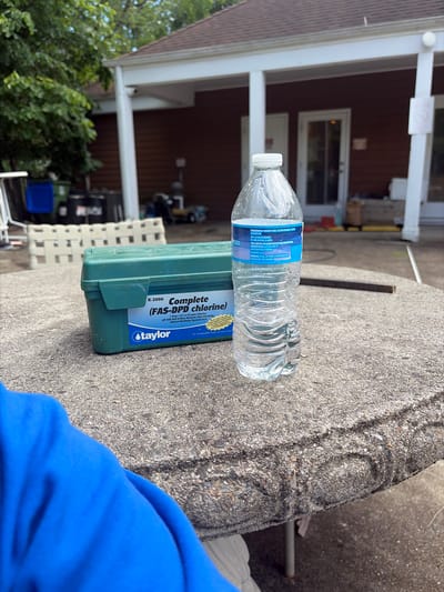 A close-up shot shows a clear plastic water bottle resting horizontally on a concrete surface, next to a green "Taylor" pool water testing kit. The kit's label reads "K-2006 Complete (FAS-DPD chlorine)". A blue article of clothing is visible in the bottom right corner.
The scene appears to be outdoors, possibly a backyard or patio setting, with a building featuring white pillars and a brown exterior visible in the background. The presence of trees and lawn furniture further suggests an outdoor residential environment. It is daytime and sunny. Based on the OCR text "1 drop 0.2 or 0.5 ppm Chlorine, pH with Acid & Base Demand, Total Alkalinity, Calcium Hardness, Cyanuric Acid", the location could be in Edison, United States of America, a suburb of New York, New Jersey, where homes typically feature pools in their backyards.