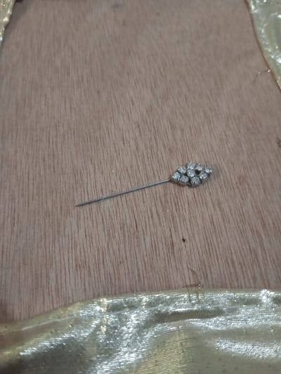 A silver-toned decorative pin rests on a light brown wooden surface. The pin features a slender shaft pointed on its left end and an elongated, marquise-shaped head on its right. The head is adorned with multiple small, clear, faceted stones, approximately 9 to 11 in number, set closely together. The wooden surface displays vertical grain patterns. In the lower portion of the image, a segment of shimmering golden fabric, exhibiting folds and a reflective texture, is visible, positioned adjacent to the wooden surface. The scene is located in Chandragiri, Nepal.