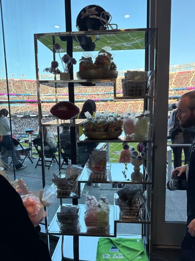 A shelf displays football memorabilia, including a helmet, football, candy baskets, and wrapped gifts. A football field filled with fans is visible through the window behind the shelf. The scene is at an elevated location, possibly a suite overlooking the field. A man with a camera appears to be taking pictures of the event.