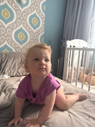 A Caucasian toddler is on a bed, on their hands and knees, wearing a purple t-shirt and diaper. The toddler has blonde curly hair and blue eyes, and is looking to the right of the frame. Behind the toddler, there is a grey tufted duvet, a pillow, a baby bottle, and a white crib with a teddy bear and pillows inside. The wall behind the bed has a pattern of grey, white, and yellow shapes. The room is lit by natural light coming from a window to the right. The location of Kondopoga, Russia is not discernable from the image.