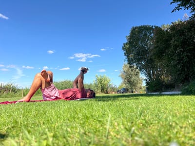 An adult male, wearing a red t-shirt and light pink shorts, lies supine on a red mat positioned on vibrant green grass. His left leg is bent with the foot flat on the mat, while his right ankle rests across his left thigh. Both arms are raised overhead, hands grasping a dark, rectangular object, possibly a mobile phone. He is barefoot and appears to be looking upwards at the object. The scene is outdoors under a clear blue sky with scattered white clouds, illuminated by direct sunlight, casting shadows. In the mid-ground, a line of dense green trees and bushes flanks the right side. A dark-colored vehicle, appearing to be an SUV, is partially visible in the distance, positioned on an unpaved path or driveway. To the left, tall green vegetation, possibly reeds or grasses, extends towards the horizon, with faint wooden posts visible. This scene is located in a rural or semi-rural area within Coruche, Portugal.