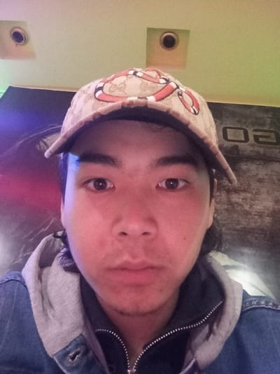 A close-up, frontal image depicts a young adult male looking directly at the camera. He wears a beige baseball cap adorned with a patterned design, including what appears to be a red, green, and white snake motif. His dark hair is visible beneath the cap and extending to the sides. He is dressed in a dark zippered garment, possibly a hoodie with a gray hood, over which he wears a blue denim jacket with visible metal buttons. His eyes are dark, and he has minimal stubble on his chin and upper lip. The indoor background is partially visible, featuring a light green ceiling with two circular, recessed fixtures. To the viewer's left, a segment of the background shows purple and blue ambient lighting. To the viewer's right, a darker surface displays partial, mirrored lettering, resembling "GO" or "CO". The specific location within Takhtakuprik, Uzbekistan, is not identifiable from the visual information presented.