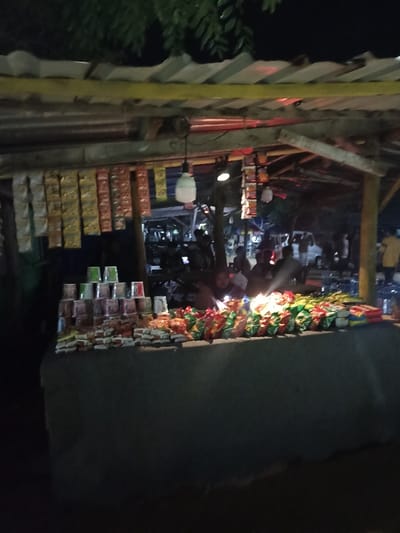 The image depicts a lively, open-air snack stall operating at night in Kecamatan Pujut, Indonesia. The stall is constructed informally with a corrugated metal roof, supported by a wooden or bamboo framework, and features a prominent yellow beam running along its front.
Artificial lighting, including a visible white bulb hanging from the roof, illuminates the stall and its offerings against the dark background of the night. Numerous individually packaged items, likely sachets of instant food or drinks, hang from the overhead structure. The main counter, draped in a dark fabric, displays a variety of products. On the left, stacks of disposable cups, some green and others with colorful patterns, are neatly arranged. The rest of the counter is covered with an abundance of packaged snacks like chips and biscuits, alongside bottles of what appear to be beverages on the far right.
Several people are visible in the scene, both behind the counter and in the background. One individual is more clearly discernible behind the counter, partially lit, seemingly attending to the stall's operations or engaging with customers. Other blurry figures and vehicles in the distance suggest a bustling roadside or market environment.
The time of day is distinctly night, creating a warm contrast between the well-lit stall and its dark surroundings. The atmosphere appears casual and active. No discernible text is visible on any of the products or stall signage.