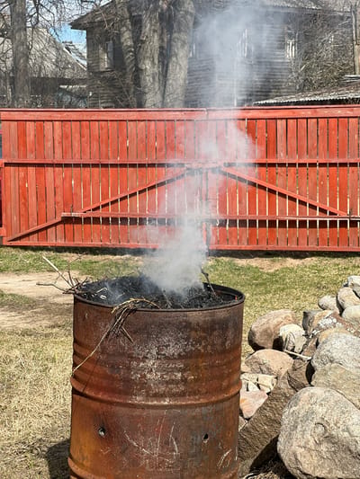 The image depicts a metal burn barrel positioned next to a red wooden fence. Smoke rises from the barrel, indicating that something is burning inside. To the right of the barrel, there is a pile of rocks. In the background, there is a building with a complex structure, potentially a house. The environment consists of grass and what appears to be early spring vegetation, suggesting a rural or suburban setting. While it is claimed that this image is from Чернышово, Russia, there is no definitive evidence in the image to confirm this.