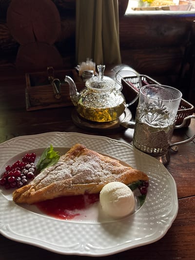 A close-up shot captures a dessert and tea setting on a rustic wooden table. In the foreground, a white, embossed plate holds a slice of strudel dusted with powdered sugar, accompanied by a scoop of vanilla ice cream, a dollop of red berry sauce, fresh red currants, and a sprig of mint. To the right, a clear glass filled with a light green beverage, possibly tea, sits in an ornate silver holder. In the background, a glass teapot filled with the same green liquid is positioned on a silver stand. Behind the teapot, a woven basket and a wooden box containing some items are visible. The scene is illuminated by warm, ambient lighting, casting soft shadows. The overall impression is one of a cozy, traditional meal or tea break. The specific location of Kamensk-Shakhtinsky, Russia, is not visually indicated in the image.