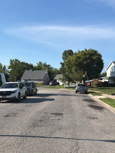 A suburban street in Levittown, New York, is depicted on a clear, sunny day. The asphalt road shows signs of wear with several patches and cracks, and a few dark, wet spots are visible. Several cars are parked along the street. In the foreground, a white SUV is parked on the left, followed by two dark SUVs. Further down the street, a dark gray SUV is parked on the right. Residential houses with manicured lawns line both sides of the street, with trees and greenery interspersed. One house, on the left, is a light gray split-level with a dark gray roof and a prominent chimney. Another house on the right is a white two-story with a distinctive arched window. A basketball hoop is visible on a pole in front of a large, leafy tree in the middle distance. The sky is a clear, bright blue with a faint white contrail stretching across it.