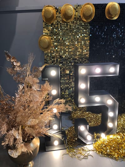 The image depicts a party decoration setup. A large black numeral "15" constructed from metal and lit with multiple bulbs stands prominently in the foreground, partially obscured by a large vase filled with golden decorative branches and foliage. The background features a sequined wall with a transition from gold to black sequins. Above the sequined wall, three gold, glitter-covered hats are suspended. The scene appears to be indoors, with a grey wall on the left and a white floor below. No people are visible in the image. This scene is likely staged for a birthday party or anniversary celebration. The city of Vaughan is known for its diverse community and vibrant event spaces, making it a plausible location for such a setup.