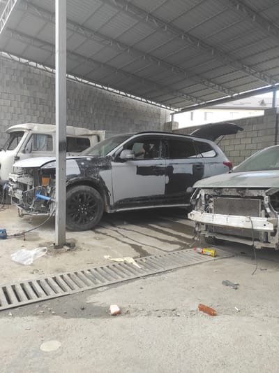 The image depicts an outdoor auto repair shop in Qairat, Kazakhstan, under a corrugated metal roof supported by a concrete wall and a metal pillar. In the foreground, a silver car with its front end disassembled, revealing the radiator and engine components, is parked on a concrete surface. To the left of this vehicle is a white utility truck, also with its front end partially dismantled. A dark gray SUV with its rear hatch open is positioned behind the utility truck, and its bodywork appears to be undergoing repair, with primer and filler visible. The ground is concrete with a metal grate drain in the middle, showing signs of wetness and scattered debris, including plastic bags, tools, and discarded items. A person is visible inside the driver's side of the gray SUV. The overall scene suggests an active but somewhat disorganized repair environment.