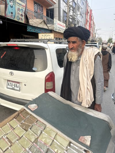The image shows an elderly man with a white beard wearing a black turban and a grey shirt, a black scarf around his neck and a beige shawl draped over his shoulders. He is standing next to a white vehicle. In the foreground, to the right of the man, is a wooden crate filled with small, individually packaged items, likely spices or herbs, arranged in rows. The man appears to be looking down or to the side.
The background shows a street scene with buildings, a variety of signs with text in an unfamiliar script, and other vehicles. The overall setting appears to be a marketplace or a busy street in a city. The lighting suggests it is daytime, possibly overcast. The image is taken from a slightly elevated angle, looking down towards the street.
Visible text includes license plates on the vehicle, with one reading "AZU 88J HONDA" and another partially obscured plate. There are also signs on the buildings, one in particular with "RAFI" visible in red lettering on the back of the vehicle. Other signs have script that is not easily decipherable. The scene suggests a bustling urban environment, possibly in Pakistan, given the mention of Chaman City Tehsil. The man's expression is difficult to discern clearly due to the angle and lighting, but he does not appear to be smiling.