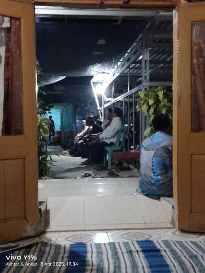 The image shows a scene taken from inside a building in Gegesik, Indonesia (latitude: -6.538, longitude: 108.43). It appears to be a residential area, possibly a home. Several people, mostly adults, are seated in a covered area outside the building. There are chairs present and the people are engaged in conversation. The environment is dimly lit with a single light source illuminating the scene. The ground is tiled and a blue and white striped mat can be seen in the foreground. The image appears to be taken through an open doorway, with the camera positioned to capture the scene from inside the building.  The doorway is wooden and partially open, allowing the viewer to look out into the outdoor gathering space.  There is a lot of greenery present.  The scene gives the impression of a casual, informal gathering.  The people are sitting in a relaxed manner and talking to each other. The image was taken in the evening as it is dark outside.
