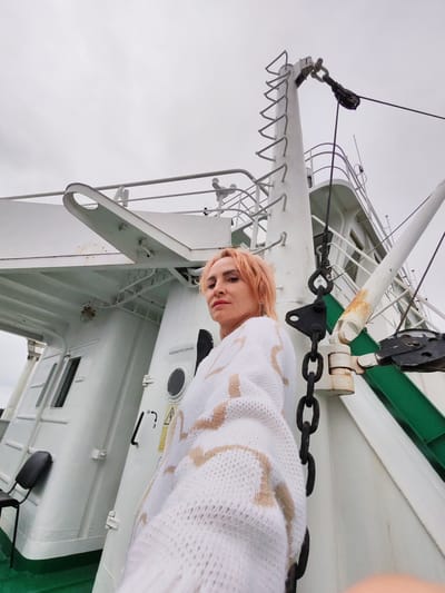 A woman with short, pink hair and light-colored makeup is posing for a selfie on the deck of a white ship. She is wearing a white knitted sweater with beige abstract patterns. Behind her, the ship's structure is visible, including a large white mast with a ladder, rigging, and metal chains. The sky is overcast and gray. The scene appears to be in an industrial or maritime setting, possibly a port in Shal'skii, Russia, indicated by the Cyrillic text "АККУМУЛЯТОРНАЯ" (Accumulator) on a door. The angle of the shot is from a low perspective, looking up at the woman and the ship's structures.