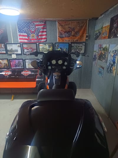 The image captures an indoor space decorated with motorcycle memorabilia. The primary focus is a black motorcycle, positioned centrally and taking up much of the frame. The seat and rear storage unit are clearly visible, obscuring some of the background elements.
Behind the motorcycle, the wall is covered with a combination of metal sheeting and decorative items related to motorcycles. Framed motorcycle images, posters, and flags are arranged along the wall. Notably, there is a flag depicting a skull wearing a motorcycle helmet with the words "BORN TO RIDE" printed on it, along with a "ROUTE 66" flag.
To the left, there is a makeshift bench constructed from oil barrels, painted black with orange seating. The lighting in the room is provided by an overhead ceiling light.
The location is identified as Surat Thani City Municipality, Thailand, based on the prompt.