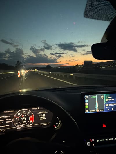 This is a first-person perspective image captured from inside a car driving on a highway at dusk. The sky displays a gradient of deep blue to orange, indicating the sun has recently set. Dark clouds are scattered across the sky, and distant city lights are visible on the horizon, suggesting the car is approaching or passing a populated area.
The car's dashboard is illuminated, showing a digital speedometer with the current speed at 71 km/h, the date and time as Monday, 15.6.2026 at 21:27, and outside temperature at 22.0ยฐC. The infotainment screen displays a navigation interface, showing a route to Sofia, Bulgaria, with an estimated arrival time of 23:30 and a distance of 180 km remaining. The car's hazard lights are on, as indicated by a red triangle symbol on the dashboard.
On the highway ahead, there are temporary traffic cones and barriers, suggesting roadwork or a lane closure. Other vehicles are visible in the distance, their headlights creating streaks of light on the wet road surface. The location is identified as being in the vicinity of Ignatievo, Bulgaria, likely on a highway leading to or from that town. No individuals are directly visible in the image.