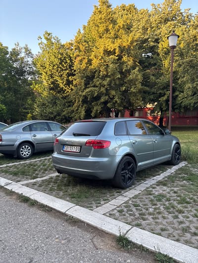The image shows two cars parked side-by-side in a parking lot paved with interlocking concrete blocks and grass. The car in the foreground is a silver Audi. Its rear is visible, showing a license plate that reads "CU 651-DO". The car behind it is also silver and appears to be a sedan.
To the left of the cars, there is a tall lamp post with a white lamp. Behind the lamp post, a large tree with lush green foliage is visible, with some branches illuminated by sunlight, creating a warm glow. The sky above is clear and blue. In the background, a building with a red facade can be seen through the trees.
The scene appears to be set outdoors during the daytime, likely late afternoon or early evening, given the warm sunlight filtering through the trees. The location is Cukarica Urban Municipality, Serbia. The overall atmosphere is peaceful and serene.