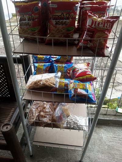 A three-tiered metal wire display rack, reinforced with cardboard on each shelf, holds various packaged snacks in what appears to be an outdoor or semi-outdoor setting.
The top shelf features several large red and orange bags of chips. Labels on these bags include "Kentang Goreng French Fries" (Indonesian for "Potato Fries French Fries"), "TOP," and "Dengan Saos" (With Sauce). One red bag clearly displays "Qtela Rasa Balado" (Balado flavor).
The middle shelf is populated with numerous smaller snack packets. Many are blue and yellow, prominently featuring "GRATIS 1 PC" (Free 1 PC) and terms like "Chocolate flavour" and "Susu" (Milk). Additionally, several clear plastic bags of individually packed snacks, possibly crackers or fried dough, and a red-and-white package labeled "ISI EXTRA" (Extra Content) are visible.
The bottom shelf also contains clear plastic bags of snacks, including more crackers or fried dough, and a bag filled with small, white, spherical snacks.
To the left of the rack, part of a wooden chair or bench is visible. The ground beneath the rack is a hard, light-colored surface, likely concrete. Beyond the immediate foreground, some green foliage indicates an outdoor environment. The lighting is diffused, suggesting it is daytime under an overcast sky. The prominent Indonesian text on the product packaging indicates these snacks are likely from an Indonesian or Southeast Asian origin.