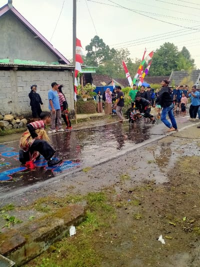 This image captures a public event on a wet, partially paved street in Tamansari, Indonesia. The ground is characterized by extensive puddles, mud, and scattered debris, including green foliage and white plastic litter. A concrete block wall with a corrugated metal roof fronts a building on the left. A utility pole with overhead wires is positioned left of center. Multiple flags, predominantly red and white Indonesian flags and multicolored decorative banners, are strung from the utility pole and across the street in the background.
Approximately 20-30 individuals are present. In the foreground left, an individual wearing a striped shirt and dark pants, with a light-colored woven wrap around their head, kneels in a puddle, interacting with colorful markings on the wet pavement. To their right, three individuals stand near the wall, observing: one in a black jacket and sunglasses, one in a light blue t-shirt, and another in a striped top.
In the mid-ground, a central activity involves two key participants. One person, dressed in black with a dark head covering, crouches over a large, ornate, circular object (possibly a traditional performance prop) that is partially submerged in a puddle. Adjacent to them, a barefoot individual in a black jacket and blue jeans, with a dark headwrap, is captured in a dynamic, backward-leaning pose with raised arms, facing the central object. A crowd of approximately 15 onlookers, dressed in casual attire, stands along the right side of the street and in the background, observing the activity. Buildings with various roof types and trees are visible under an overcast sky in the distant background.