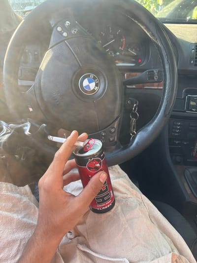 A first-person perspective shot inside a car, focusing on a hand holding a lit cigarette and a can of energy drink. The hand, with visible hair on the forearm, is positioned in the lower-right quadrant of the frame. The cigarette, held between the thumb and index finger, emits a trail of smoke that drifts to the left. Next to the cigarette, a red and black can of "REDBULL ENERGY DRINK CLASSIC" is held by the same hand. The background is dominated by the steering wheel of a BMW, featuring the iconic circular logo. Control buttons are visible on the steering wheel, and part of the dashboard with gauges is also in view. The interior of the car appears to be well-lit by sunlight, casting a bright glow across the scene. The car's interior is partially visible, with wooden trim accents and dark plastic surfaces. The focus is tightly on the hand, cigarette, and can, with the car's interior serving as a contextual backdrop. The location is identified as Letnitsa, Bulgaria.